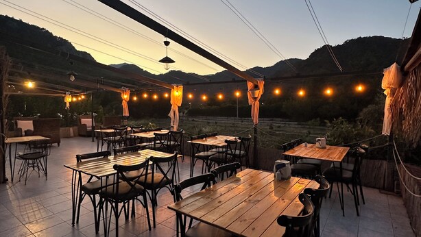 An outdoor patio with wooden tables and chairs, string lights, and a view of hills in the background. - Gaya Pansiyon (Kemer)