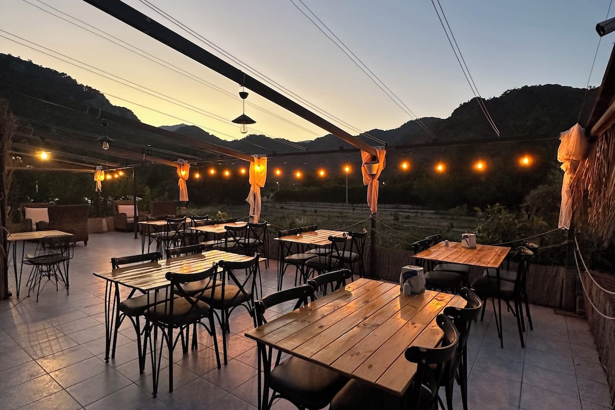 Een terras buiten met houten tafels en stoelen, lichtjes en een uitzicht op heuvels.