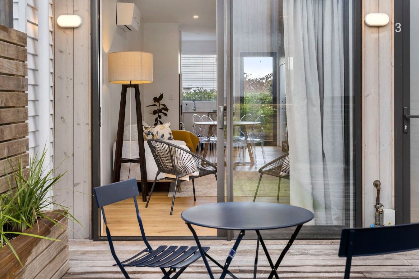 A modern outdoor seating area with a table and chairs, a lamp, and a view of the interior through a glass door.