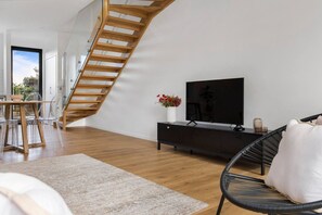 A modern living room with a wooden staircase, a flat-screen TV, a dining area with a glass table and chairs, and a black sofa.