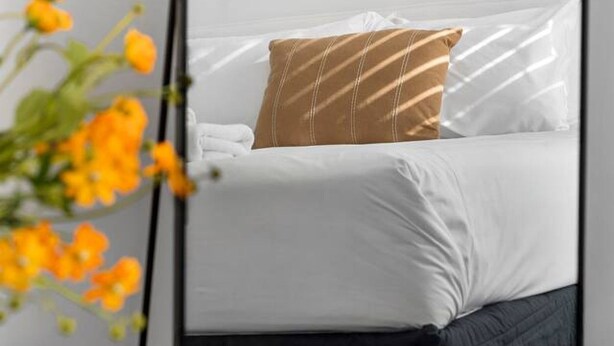 A bed with white bedding and a brown striped pillow, a small bedside table, and an air conditioning unit mounted on the wall.