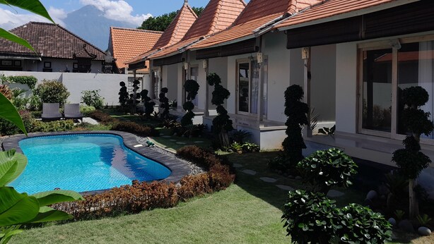 Un área junto a la piscina con un jardín y varias plantas, y un edificio con múltiples tejados.