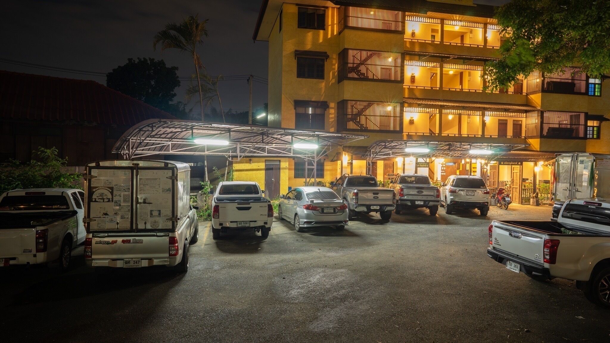 A parking lot with several vehicles, including trucks and cars, parked in front of a building with multiple balconies and illuminated windows.