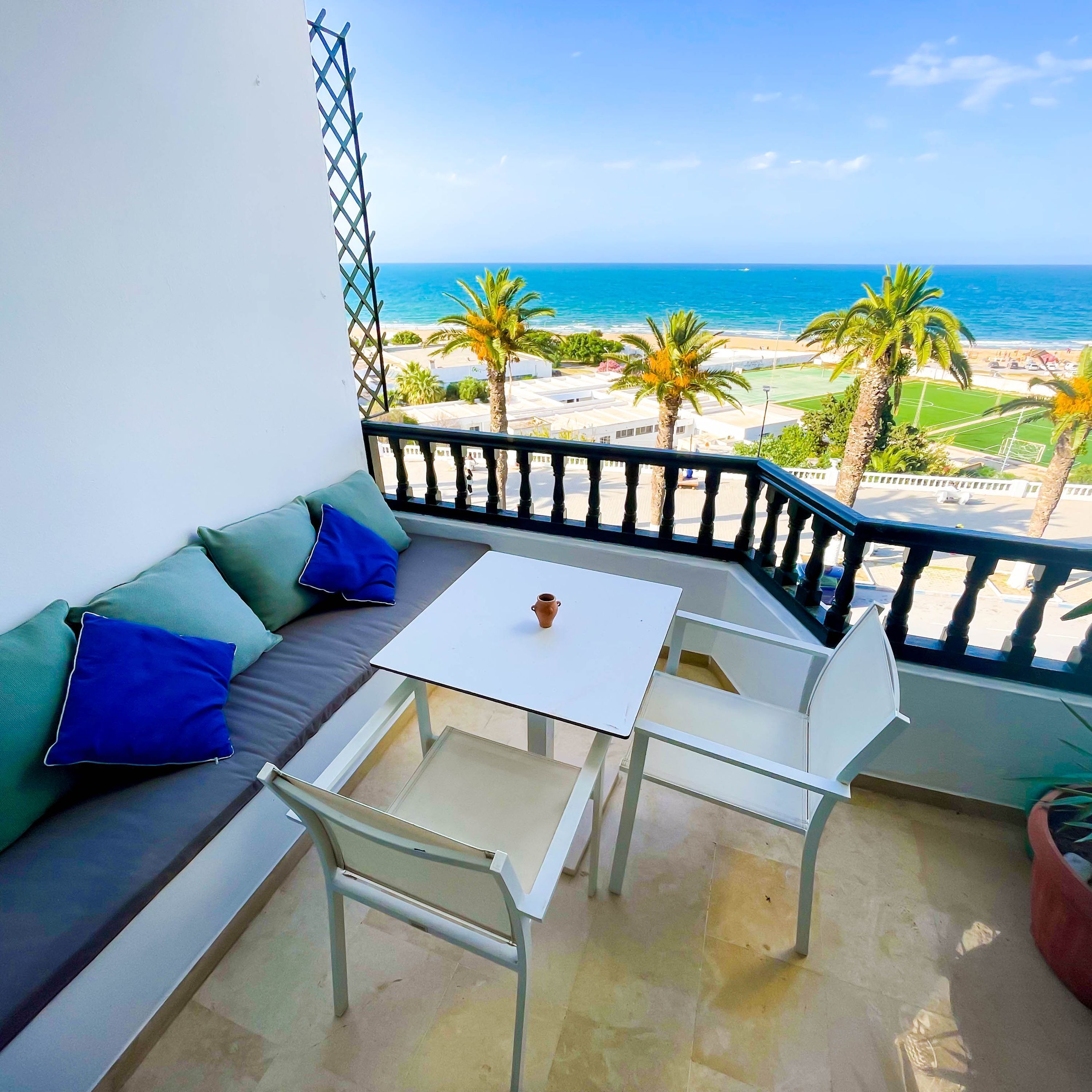 Un balcon avec une table blanche, un canapé vert agrémenté de coussins bleus, et une vue sur la mer et les palmiers.