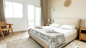 Un dormitorio bien iluminado con una cama grande, una silla de madera, una mesita y vistas al exterior por un ventanal corredero.