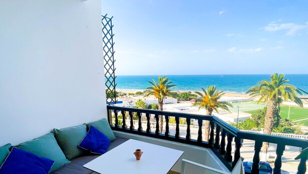 Un balcón con una mesa y sillas blancas, un sofá con cojines, y vistas a la playa y al océano.