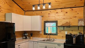 Une cuisine avec des murs en bois, des meubles blancs, un réfrigérateur noir, un évier et une fenêtre avec des stores.