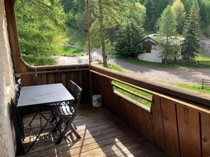 A wooden deck with a table and chairs, overlooking a forested area and a small building.