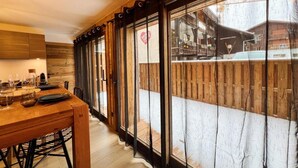 Une table à manger en bois avec des chaises, un coin cuisine avec des meubles en bois, et une vue sur un espace extérieur enneigé, visible à travers de grandes fenêtres.