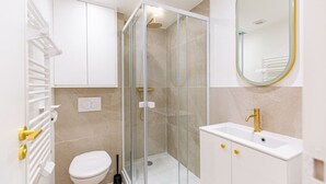 A modern bathroom with a glass-enclosed shower, a white toilet, a white sink with gold fixtures, and a large mirror. - Lavie Maison: Studio Équipé (Bordeaux)