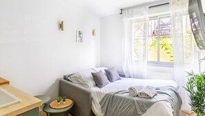 A modern bedroom with a bed, bedside table, and a window with sheer curtains. - Lavie Maison: Studio Équipé (Bordeaux)