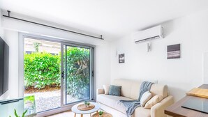 A modern living room with a sofa, a coffee table, and a TV. There is a sliding glass door leading to a garden.
