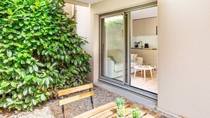 A patio with a wooden bench, a green bush, and a sliding glass door leading to a modern kitchen and living area.