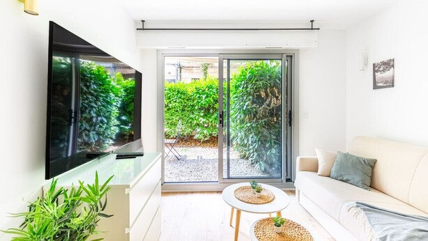 A modern living room with a flat-screen TV, a sofa, a small table, and a sliding glass door leading to a garden.