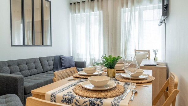 A dining area with a wooden table set for four, surrounded by grey sofas and a small wooden side table. - Lavie Maison A/C & Balneo (Paris)