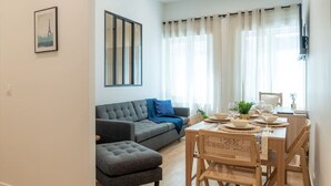 A modern living room with a grey sofa, a dining table set for four, and a large window with sheer curtains. - Lavie Maison A/C & Balneo (Paris)