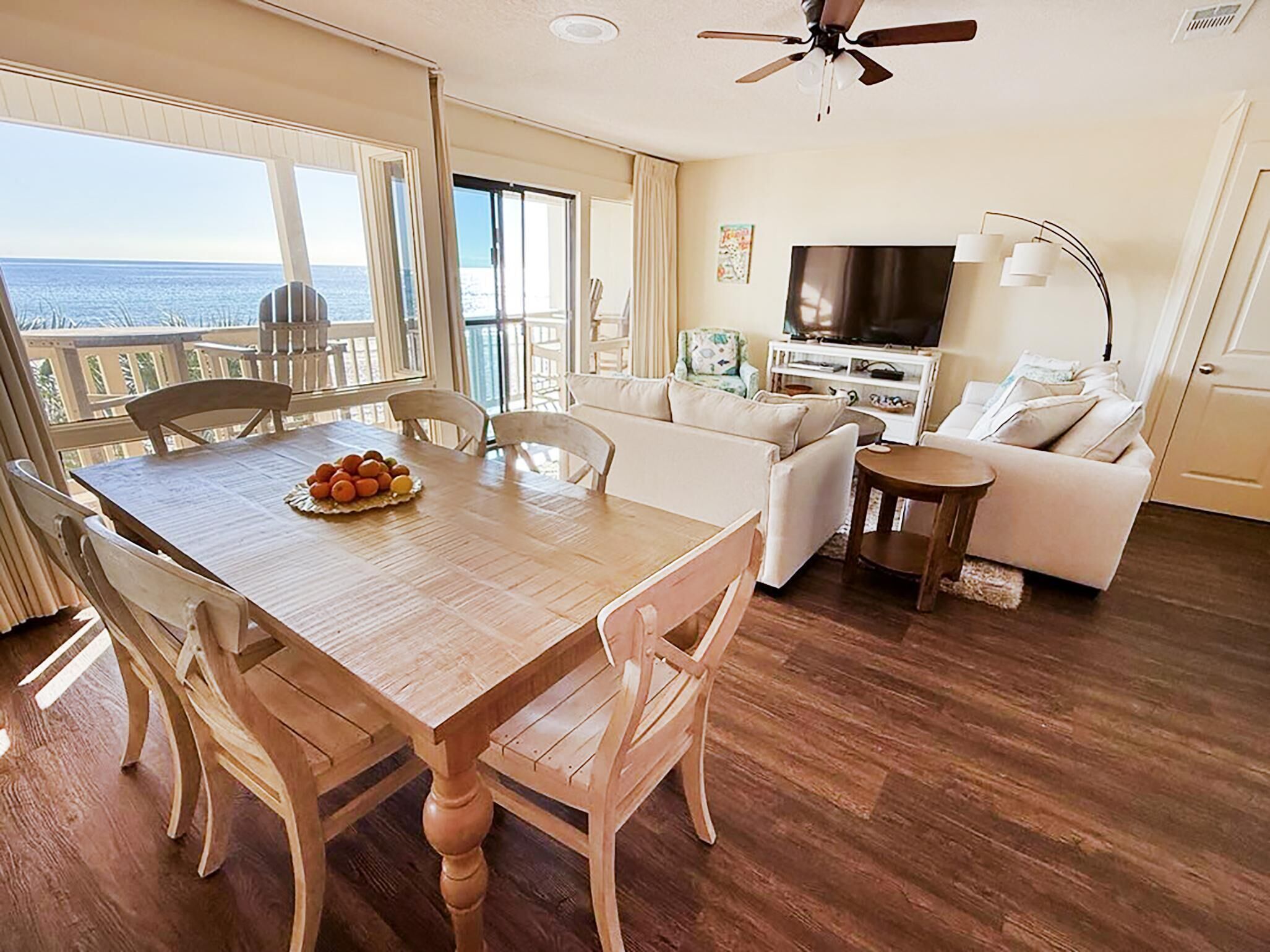 Un coin repas avec une table et des chaises en bois, un canapé blanc, un téléviseur et un balcon avec vue sur l’océan.