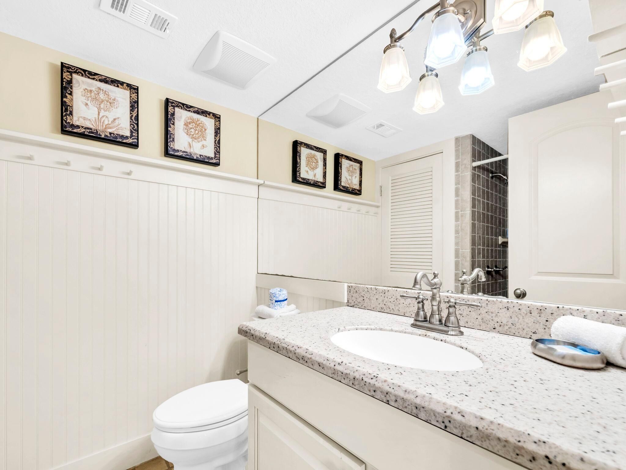 Un baño con un inodoro blanco, un lavamanos con encimera salpicada y tres cuadros con motivos florales en la pared.