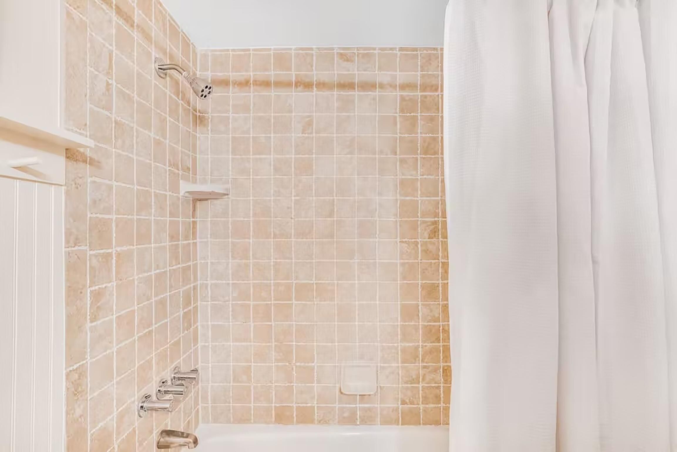 Un baño con paredes de baldosas color beige, una bañera blanca y una ducha con cortina.