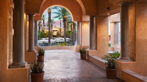 A covered walkway with columns, potted plants, and a view of outdoor seating and palm trees.