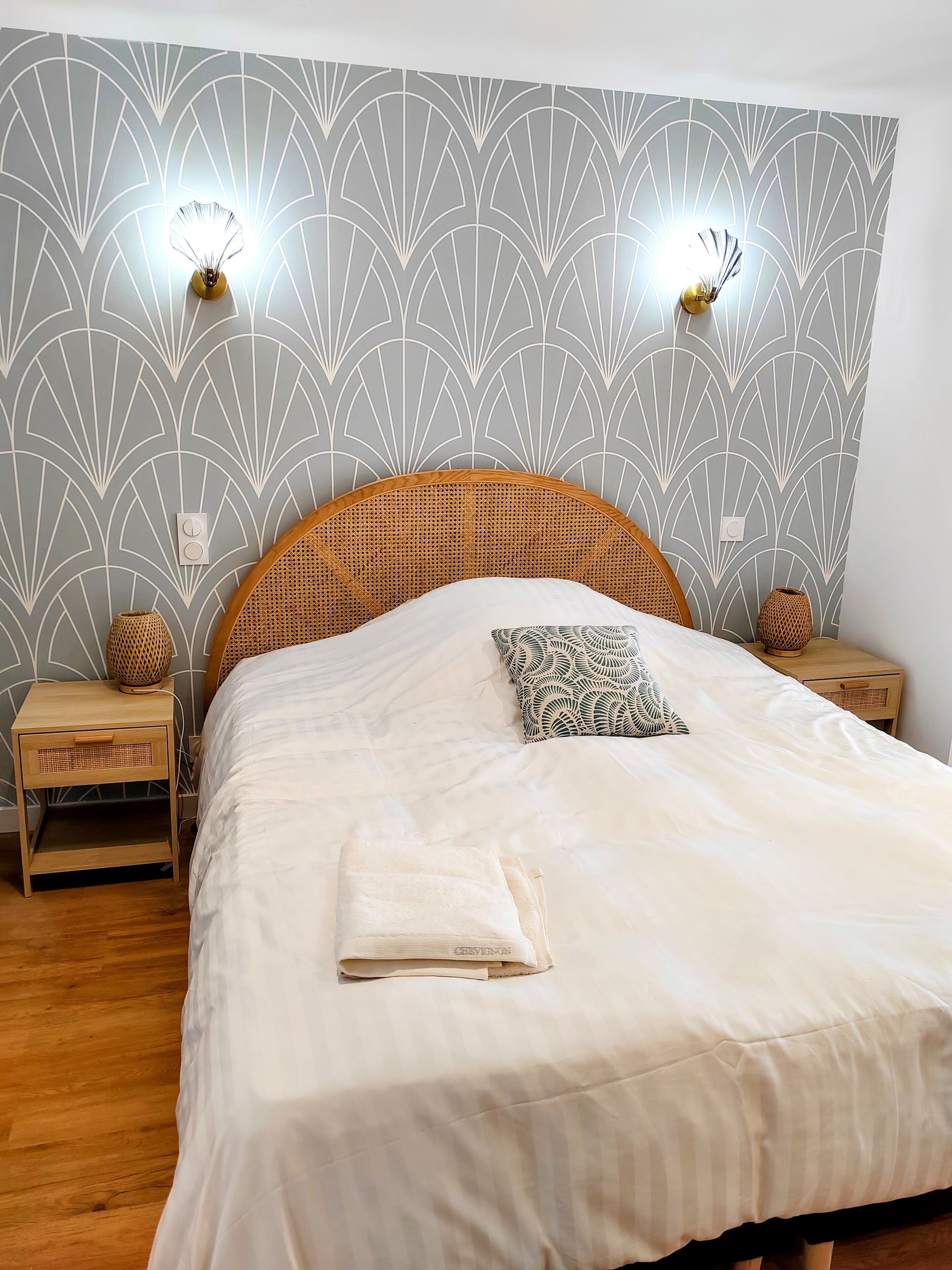 A neatly made bed with a white comforter and a folded towel, flanked by two bedside tables with woven baskets, against a wall with a geometric pattern.