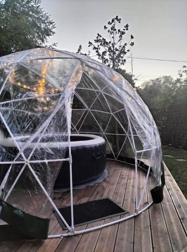 Une structure en forme de dôme géodésique abritant un jacuzzi, le tout entouré d’une enveloppe transparente et installé sur une terrasse en bois.