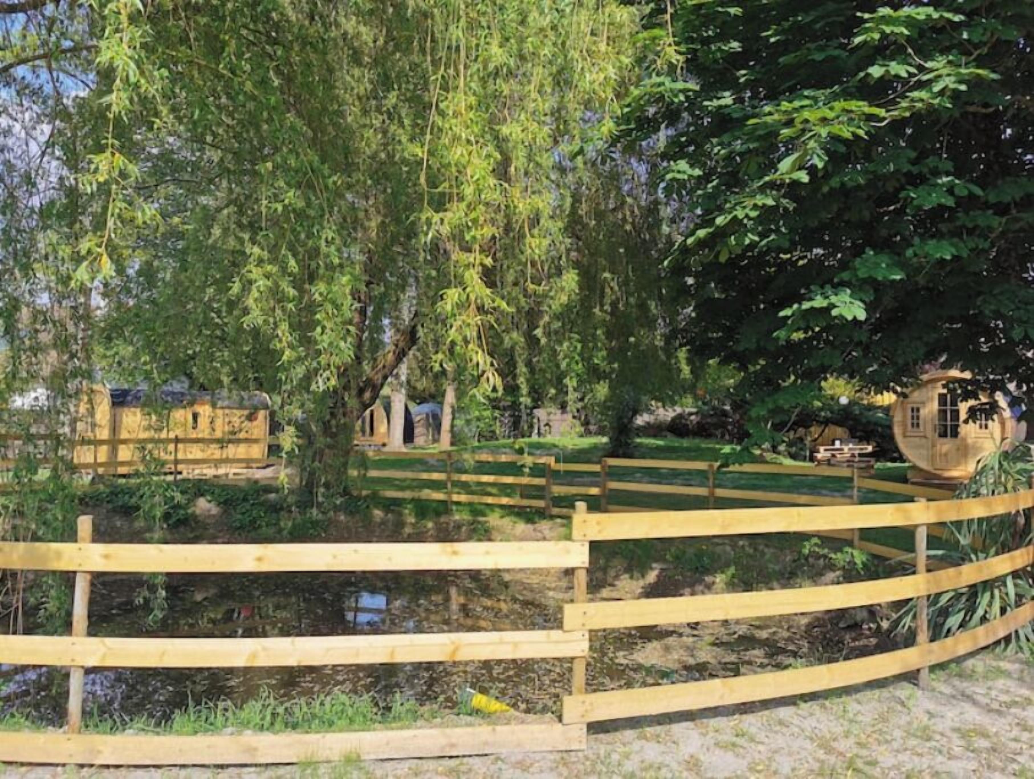 Une clôture en bois entoure un étang, avec des arbres et de petites constructions en arrière-plan.