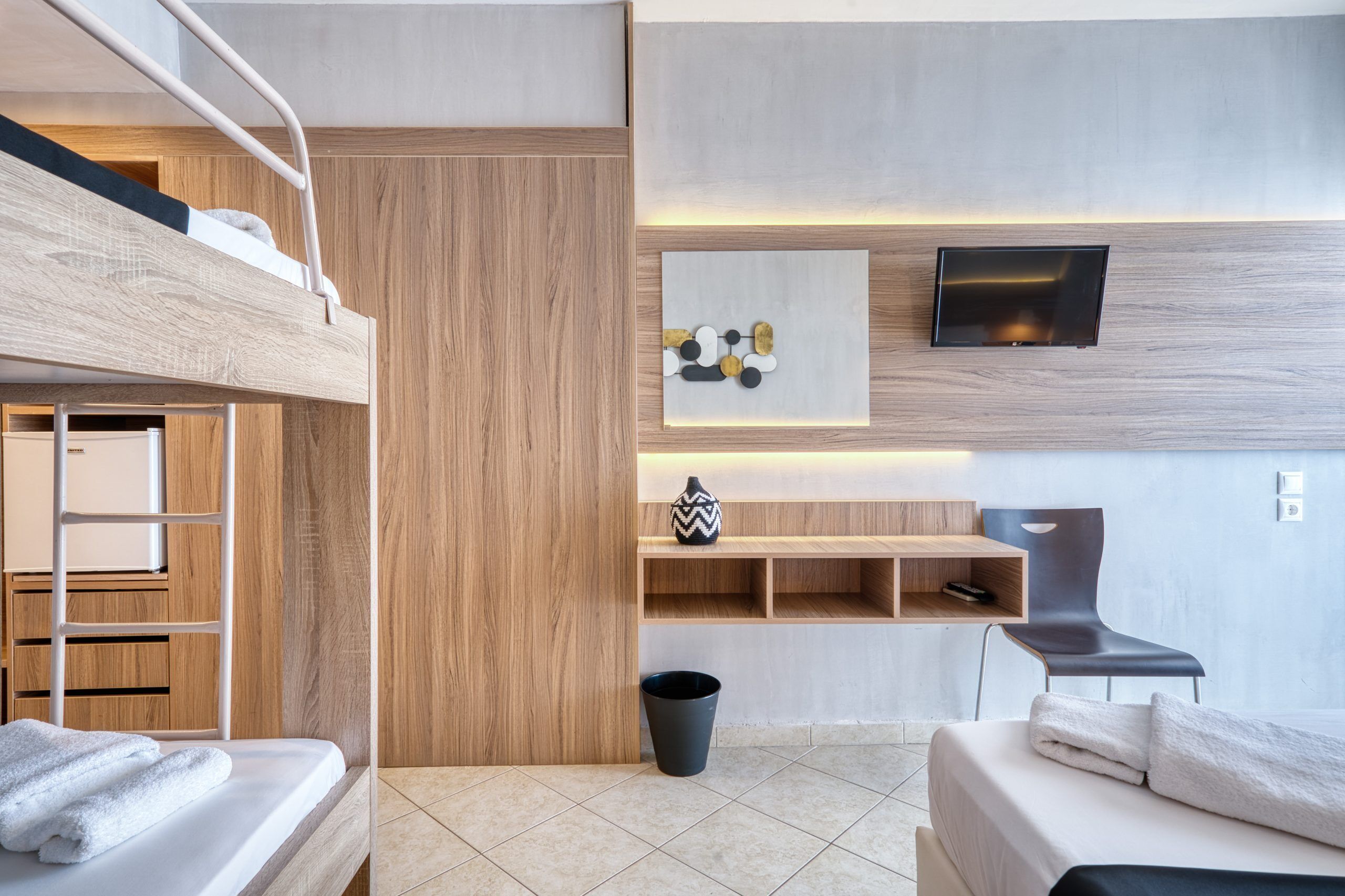 Een moderne hotelkamer met een stapelbed, een flatscreen-tv, een klein plankje met decoratieve voorwerpen en een stoel.
