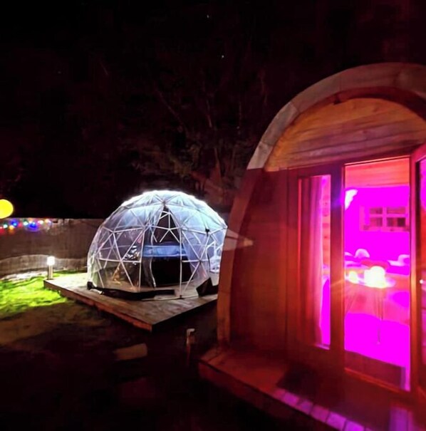 A geodesic dome structure with a wooden platform and a pink-lit window.