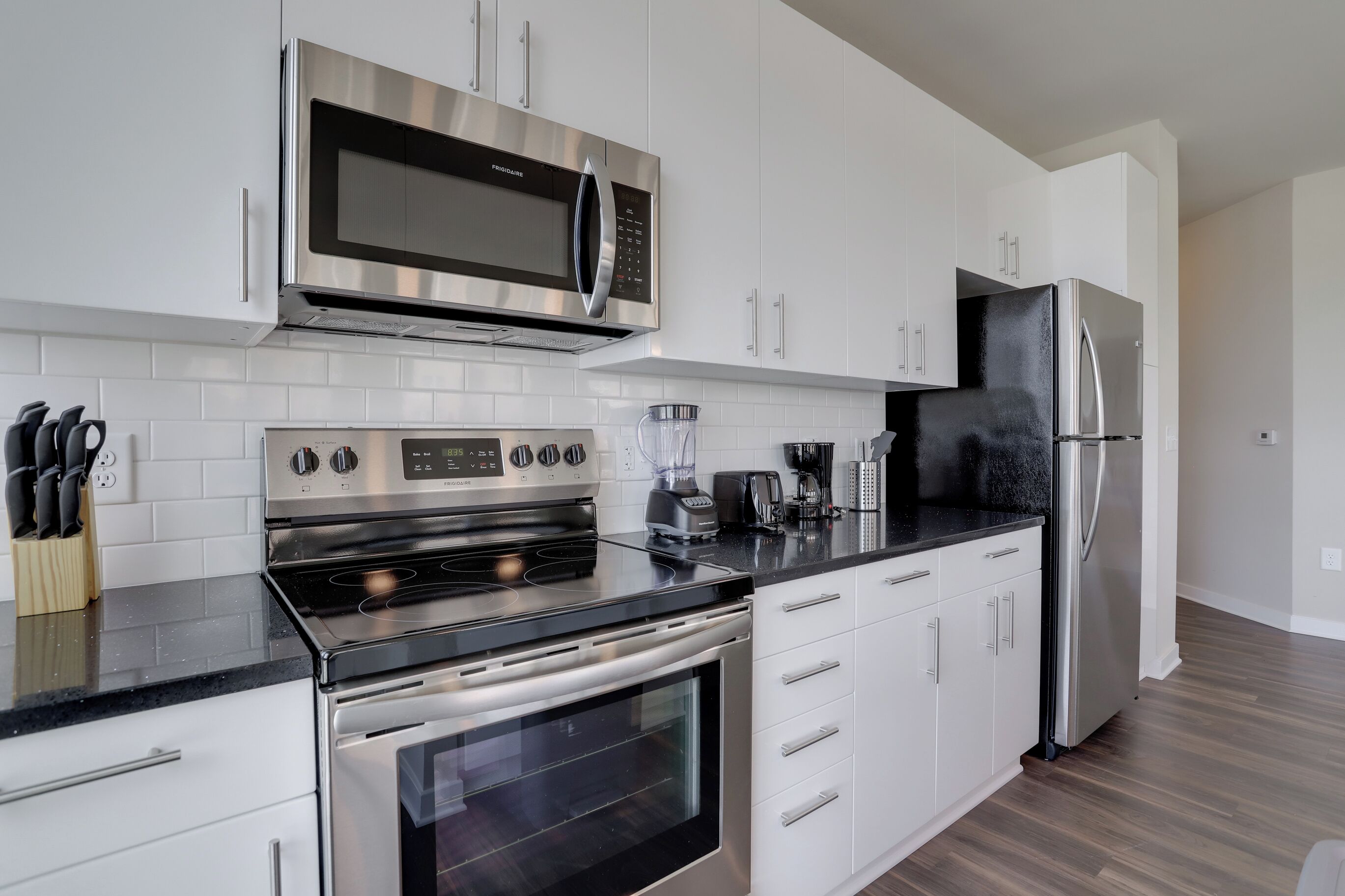 A modern kitchen with white cabinets, stainless steel appliances, a black countertop, and a wooden floor.