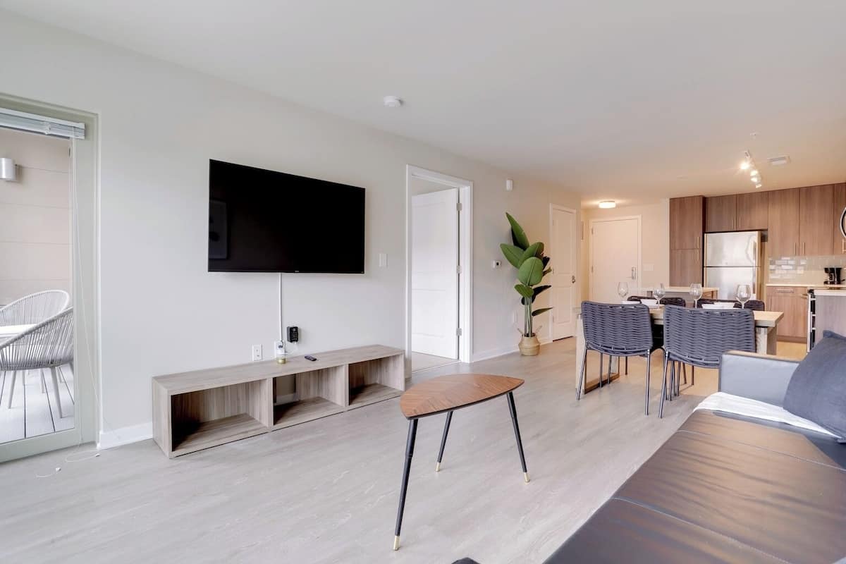 A modern living room with a flat-screen TV, a wooden bench, a dining area with wicker chairs, and a kitchen in the background.