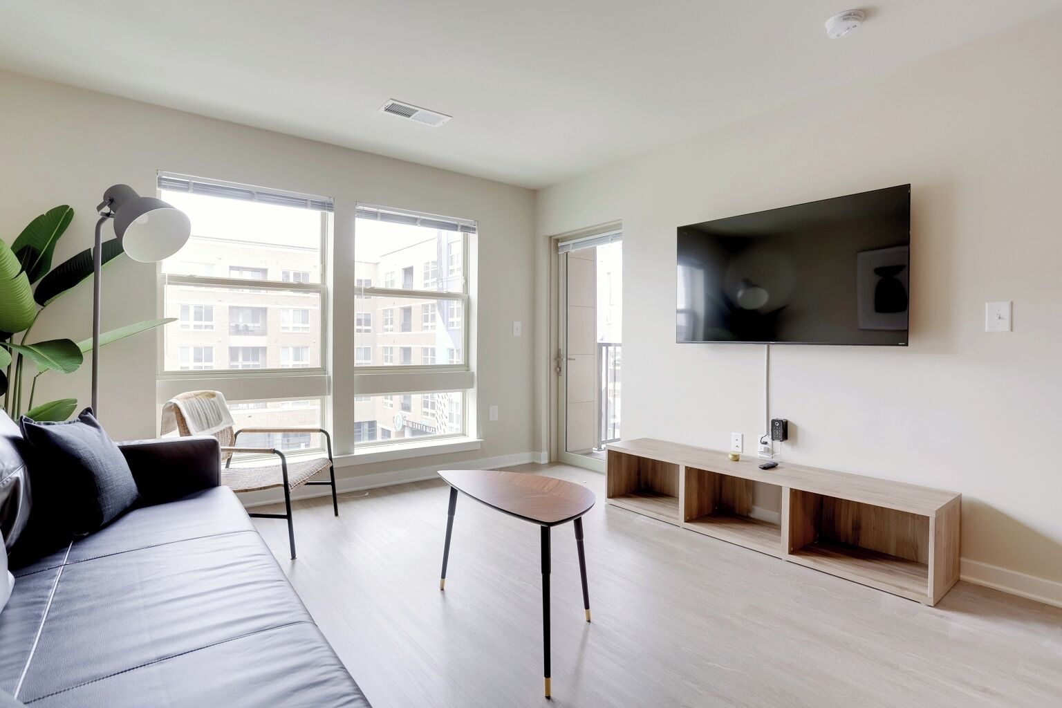 A modern living room with a sofa, a coffee table, a TV on a wooden stand, and large windows with a view of buildings.