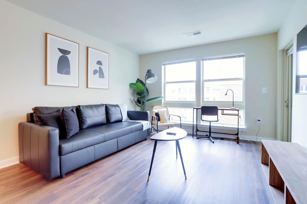 A modern living room with a grey sofa, a small round table, and a chair. There are two framed pictures on the wall, a large window, and a plant.