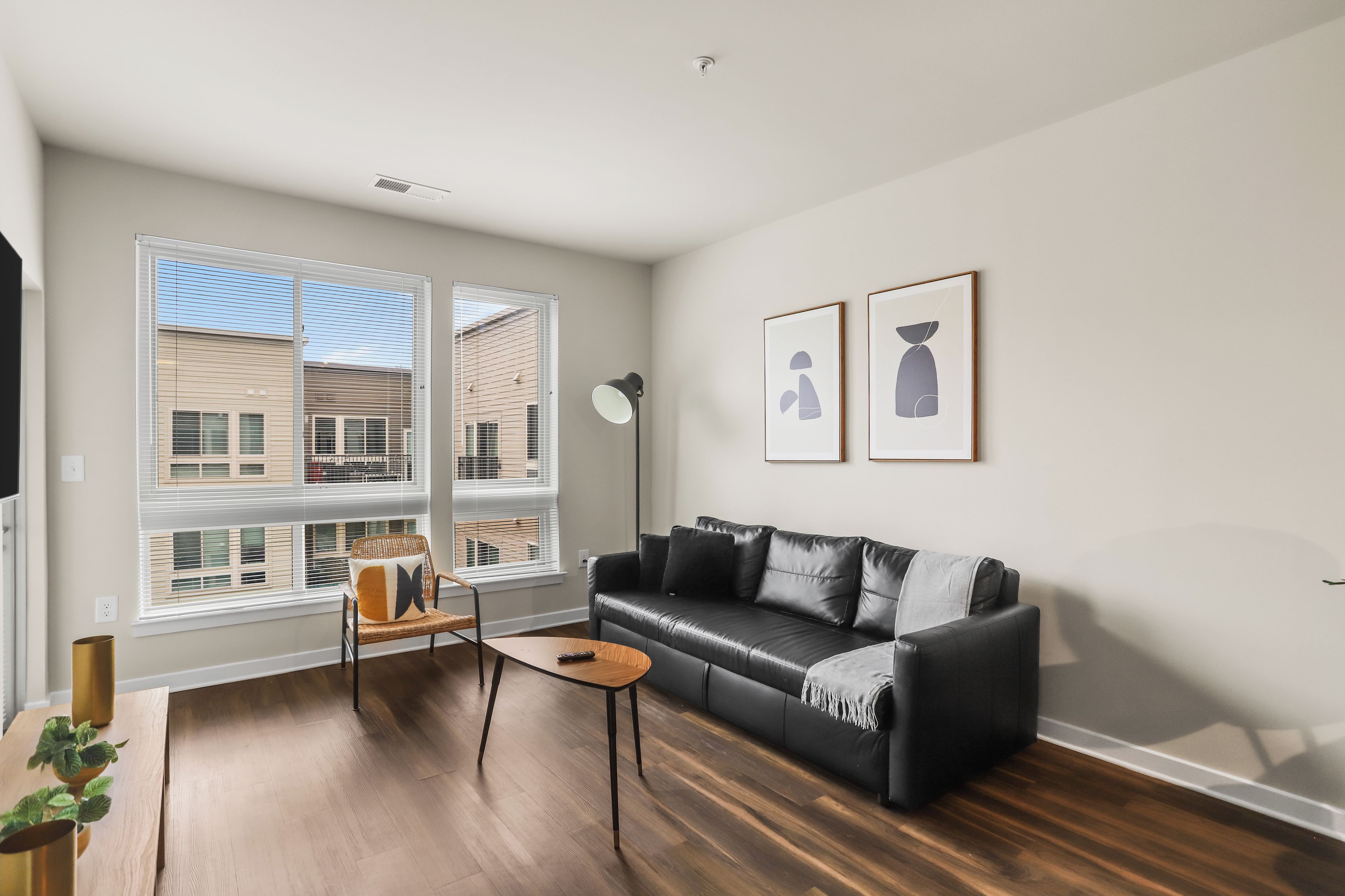 A modern living room with a black leather sofa, a wooden coffee table, a wicker chair, and two large windows with blinds.