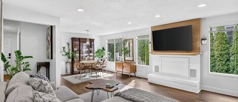 A modern living room with a large sofa, a dining area with a table and chairs, a television mounted on a wooden wall, and a fireplace.