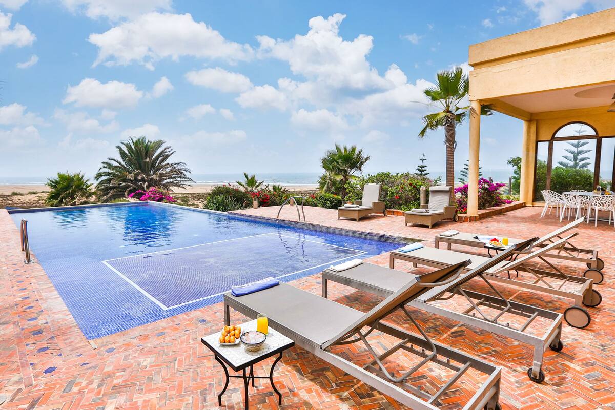 A pool area with lounge chairs and a table, surrounded by palm trees and a view of the beach.