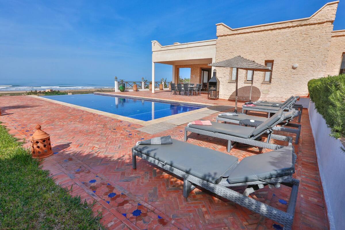 A pool area with lounge chairs, a barbecue, and a view of the ocean.