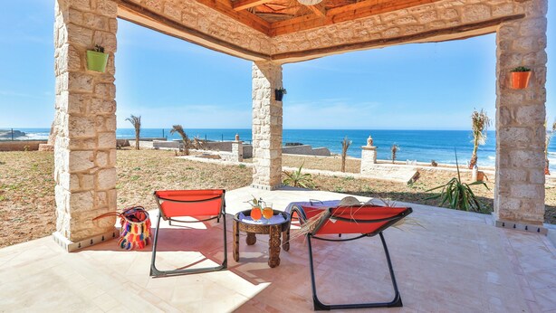 Ein überdachter Außenbereich mit Steinsäulen, einem Tisch mit Getränken und Blick auf Strand und Meer.