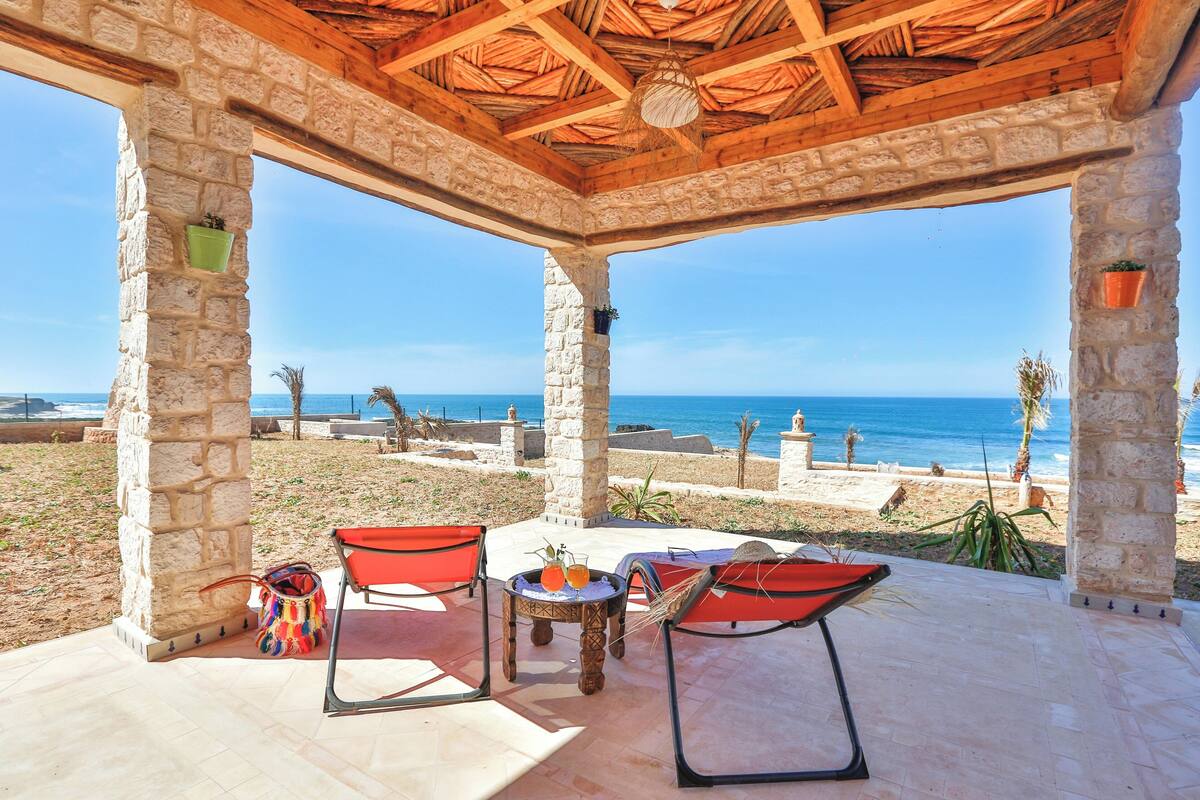 A covered outdoor seating area with stone pillars, a table with drinks, and a view of the beach and ocean.
