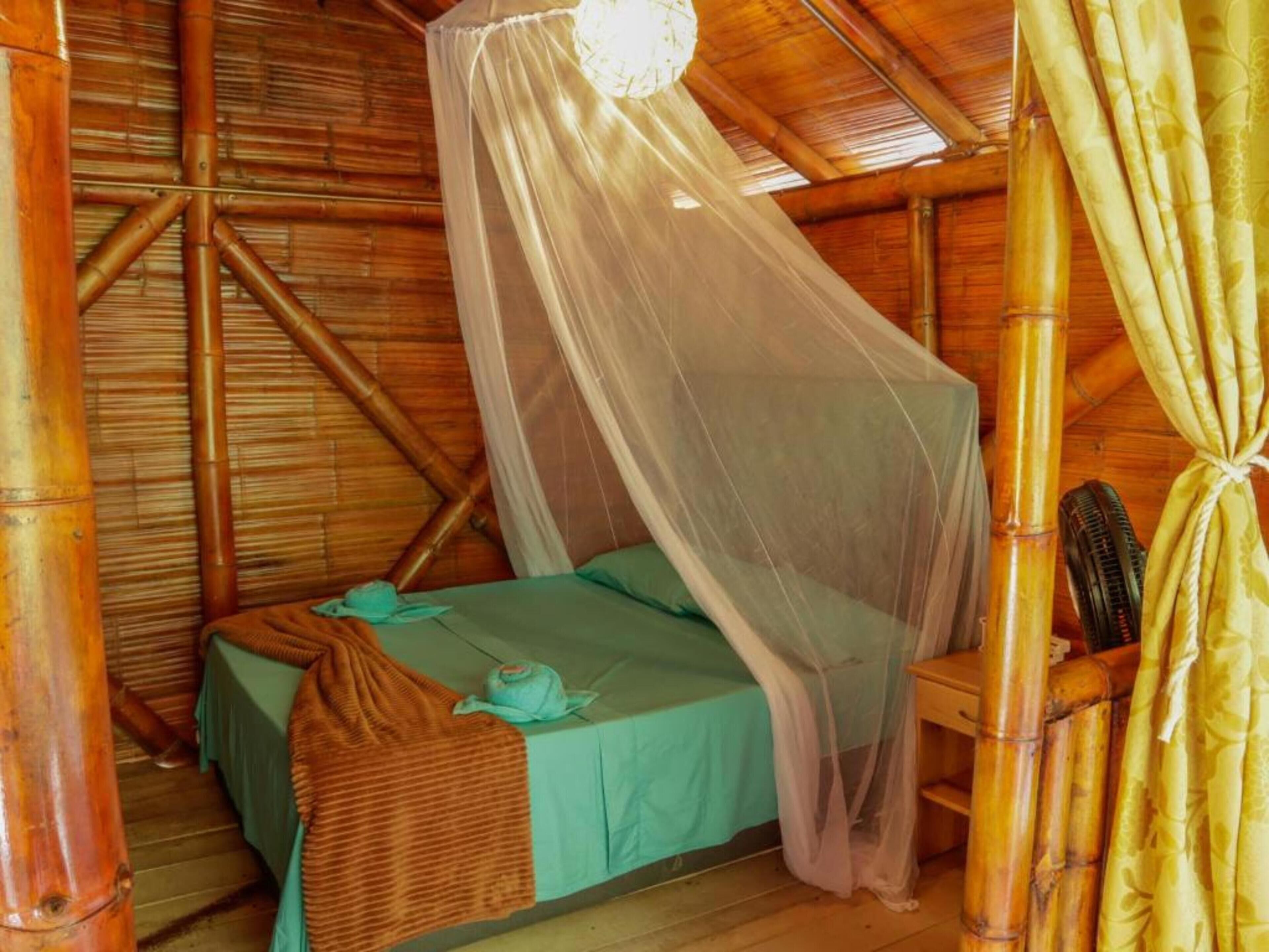 A single bed with a mosquito net, a wooden chair, and a bamboo fan in a room with bamboo walls and a sloped ceiling.