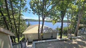 A house with a gravel driveway and a wooden deck, surrounded by trees and overlooking a body of water. - Ozarks Lake House, Private Dock w/ Boat and Jetski lifts! (Rocky Mount)