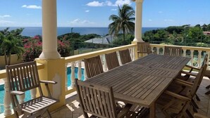 Un espace extérieur abrité, doté d’une table et de chaises en bois, d’une piscine et offrant une vue sur l’océan et les palmiers.
