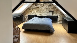 A bedroom with a sloped ceiling, stone wall, wooden floor, a bed, a dresser, and a hanging light fixture. - Maison Entièrement Rénovée Avec Billard en Plein Cœur du Légué et à 5 min Plage (Plérin)