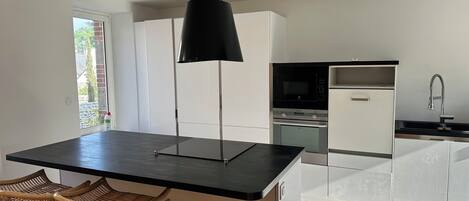 A modern kitchen with a black island, white cabinets, a built-in oven, and a stainless steel sink. There are two wicker chairs at the island.