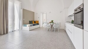 A modern kitchen with white cabinets and a stainless steel oven. There is a dining area with a white table and chairs. A white sofa with colorful cushions is in the background. - La Piccola Corte - Appartamento1 by BarbarHouse (Maruggio)