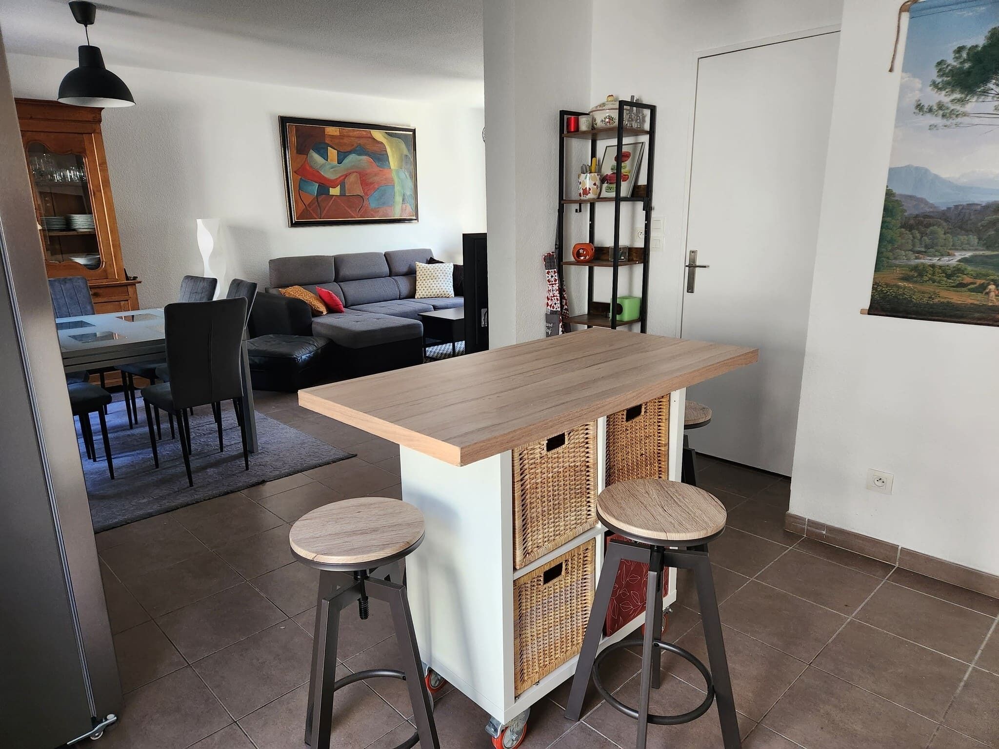 Una cocina moderna con una isla de madera, dos taburetes, un comedor con mesa de vidrio y una sala de estar con sofá y un cuadro.
