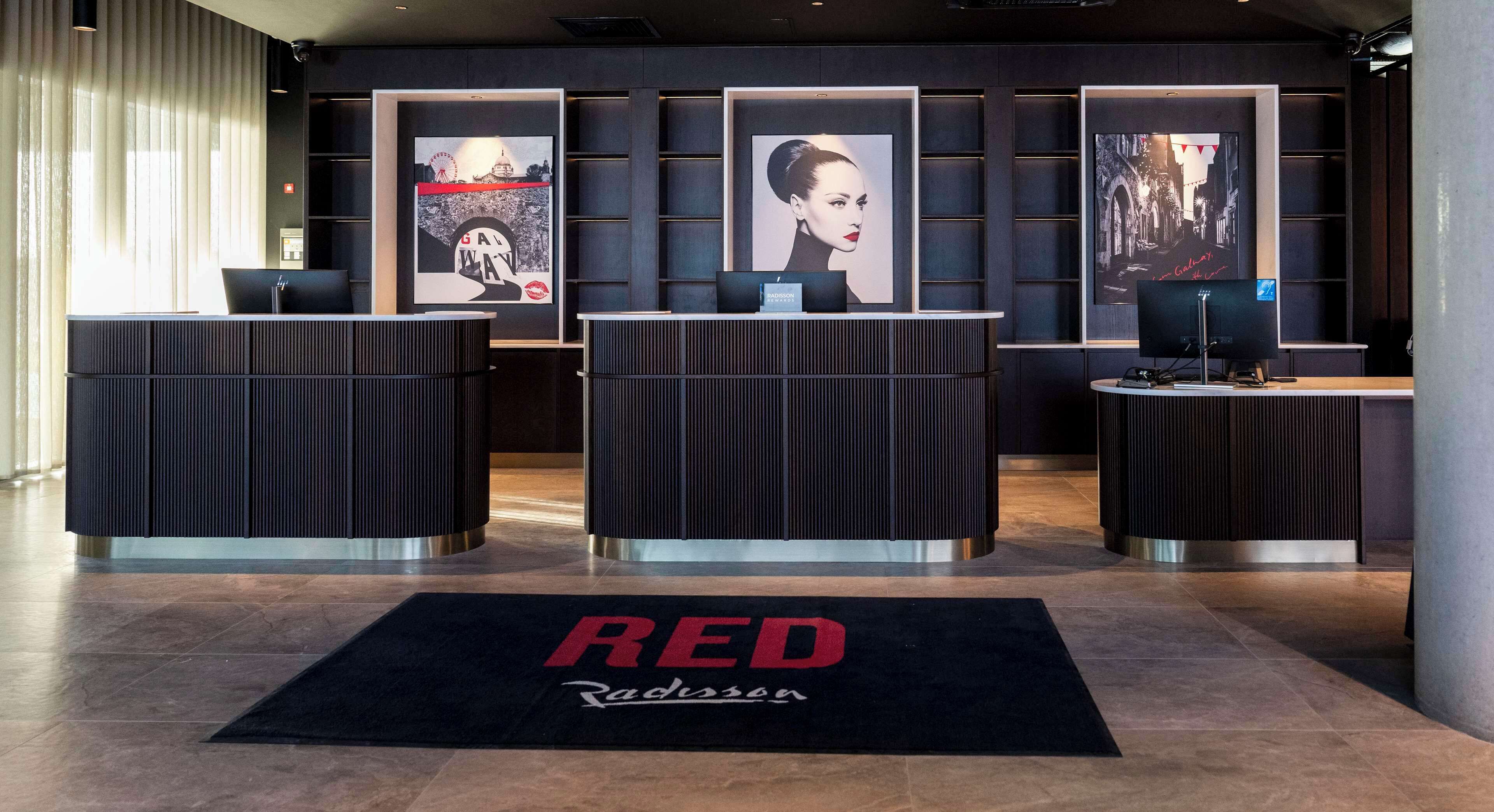 Un área de recepción con tres mostradores negros, una alfombra negra y roja con el texto 'RED Radisson' y obras de arte enmarcadas en las paredes.