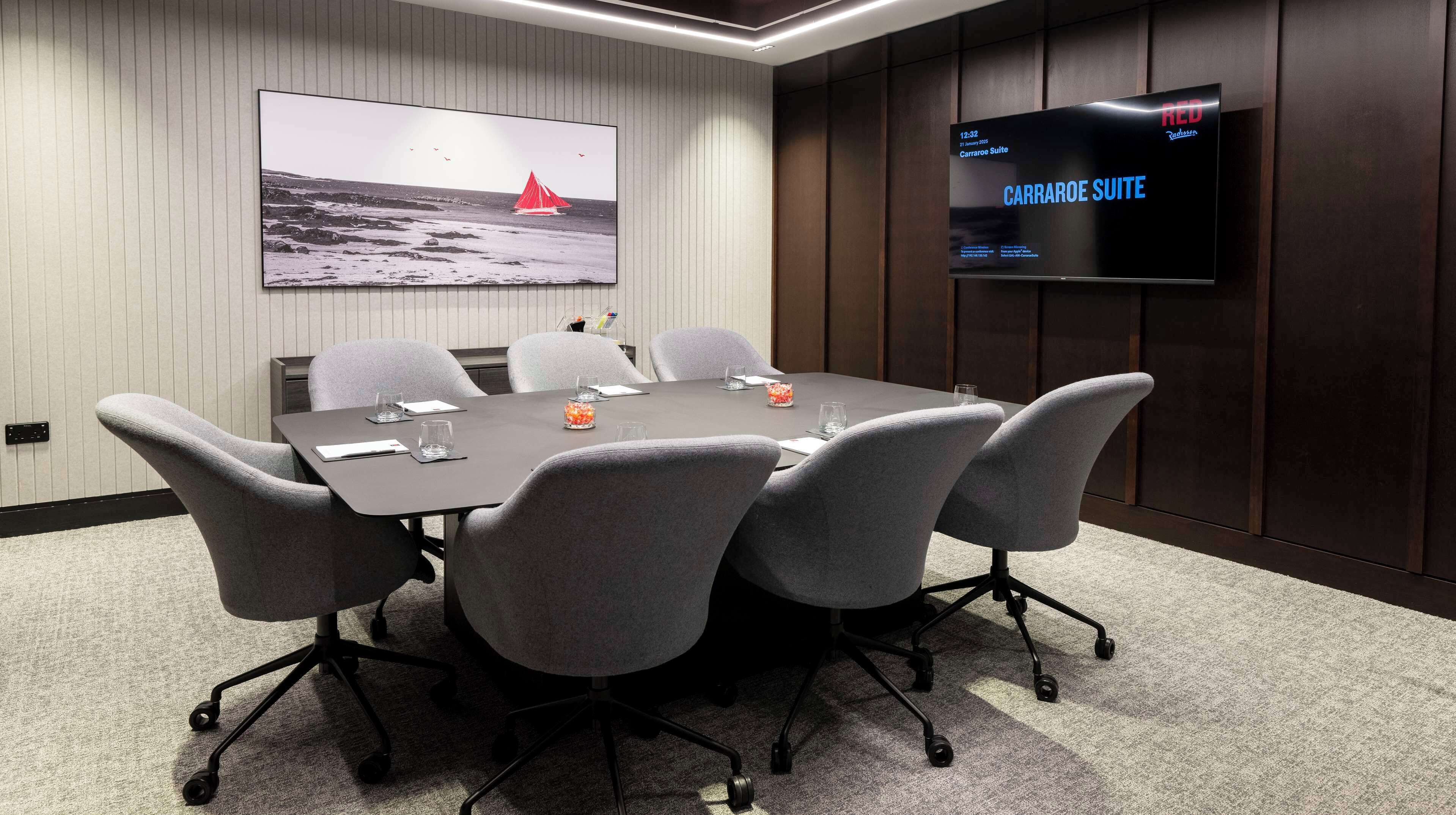 A conference room with a large table, six chairs, a flat-screen TV displaying 'CARRAROES SUITE', and a framed picture of a sailboat on the wall.
