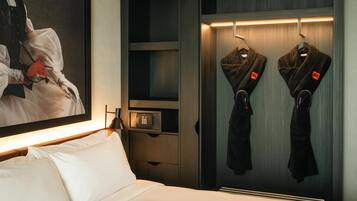 A hotel room with a bed, a painting, a coat rack with two coats, and a safe.