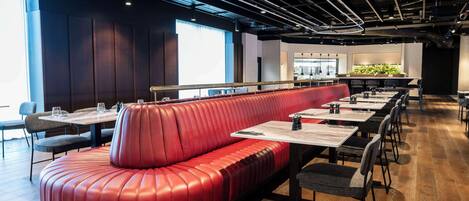 A modern restaurant with a red leather booth, wooden tables, and a large window providing natural light.